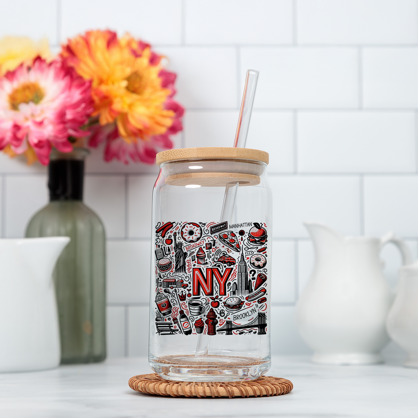 A glass jar with a straw, featuring a label with the word "NY" and various icons, is placed on a wicker coaster in a kitchen setting with a white tiled wall and colorful flowers in the background.