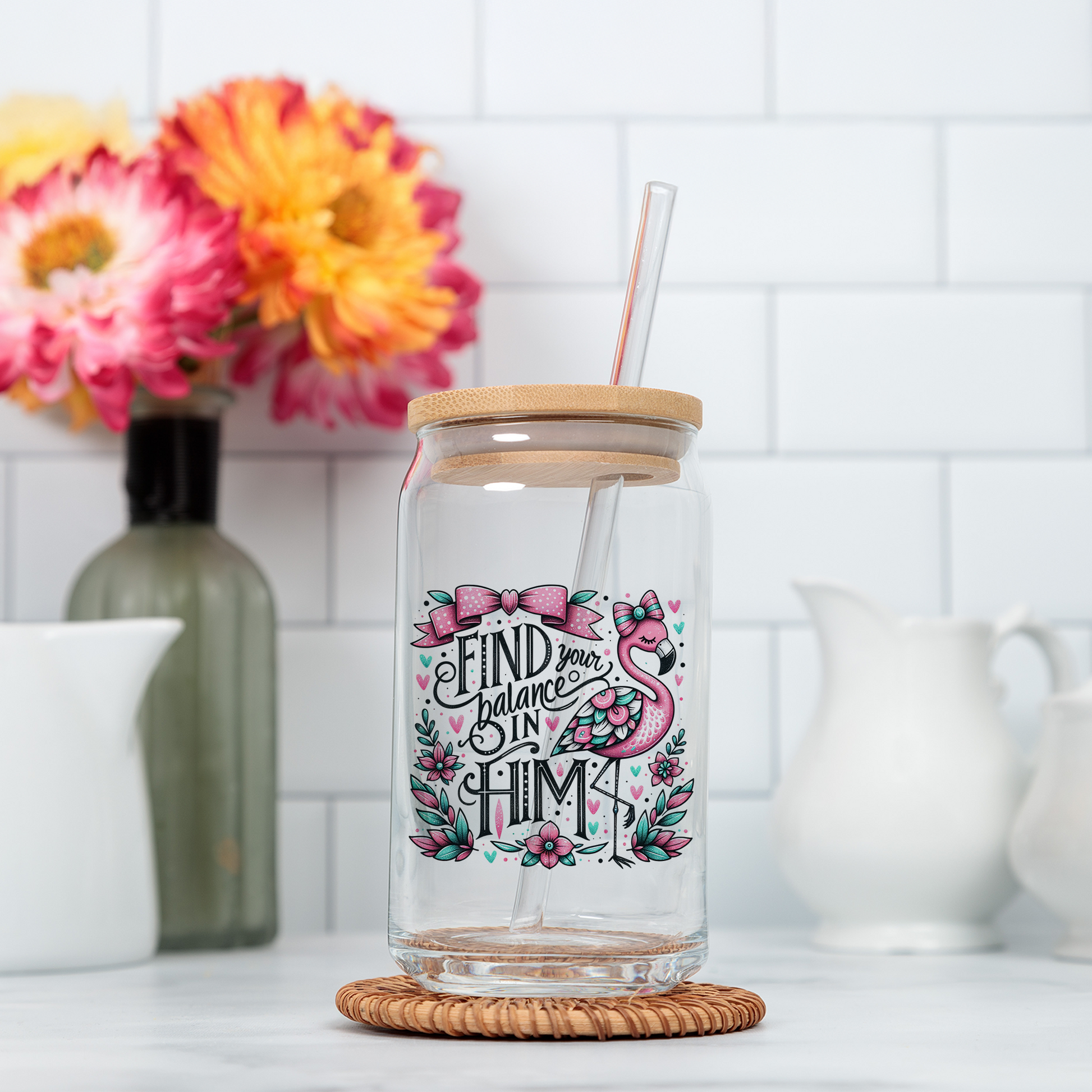A glass jar with a straw, featuring a colorful design and text, sits on a wicker coaster in a kitchen setting with a vase of flowers and other kitchen items in the background.