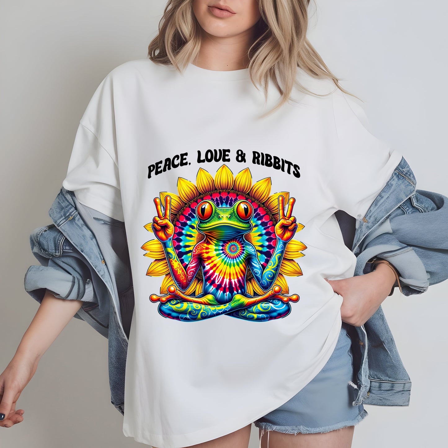 A woman wearing a white t-shirt with a colorful tie-dye design of a frog holding a peace sign, with the text "Peace. Love. RibBITS" printed on it.