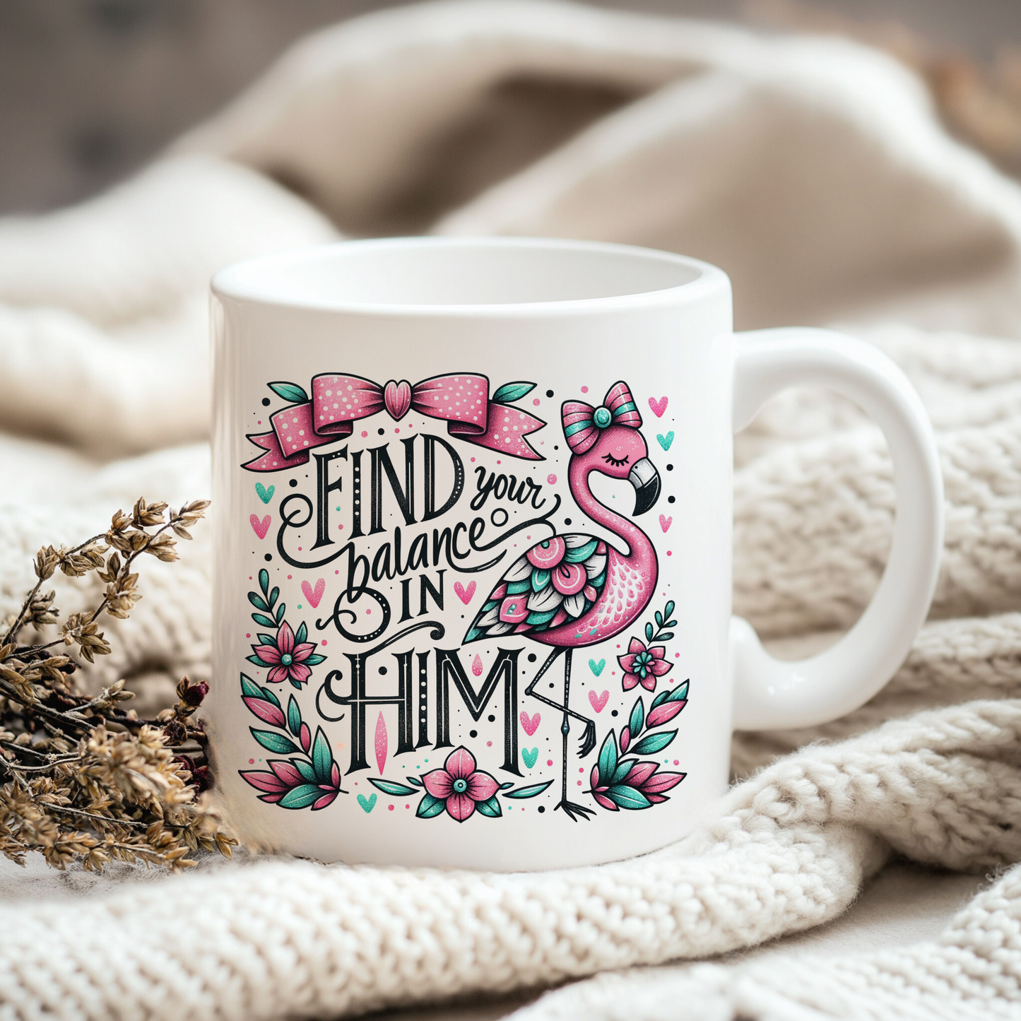 A white ceramic mug with a colorful design featuring a pink flamingo and the text "Find your balance in Him" is placed on a white blanket.