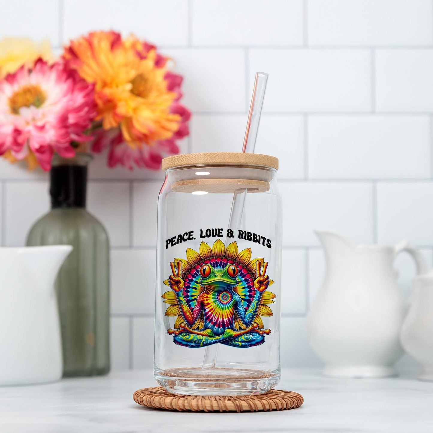 A glass jar with a colorful frog design and the text "Peace, Love & RibBITS" printed on it, placed on a woven coaster.